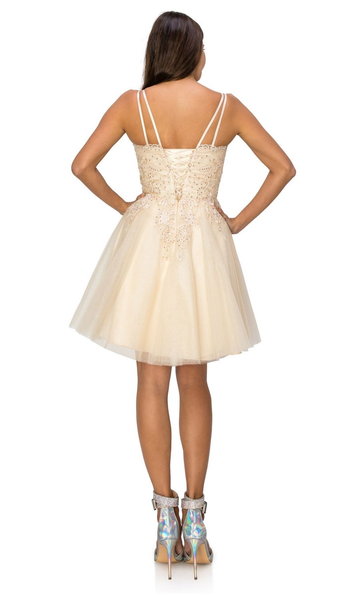 A woman stands with her back to the camera, wearing the Corset-Back Short Glitter Homecoming Dress 5125J in light beige, paired with silver holographic ankle boots.